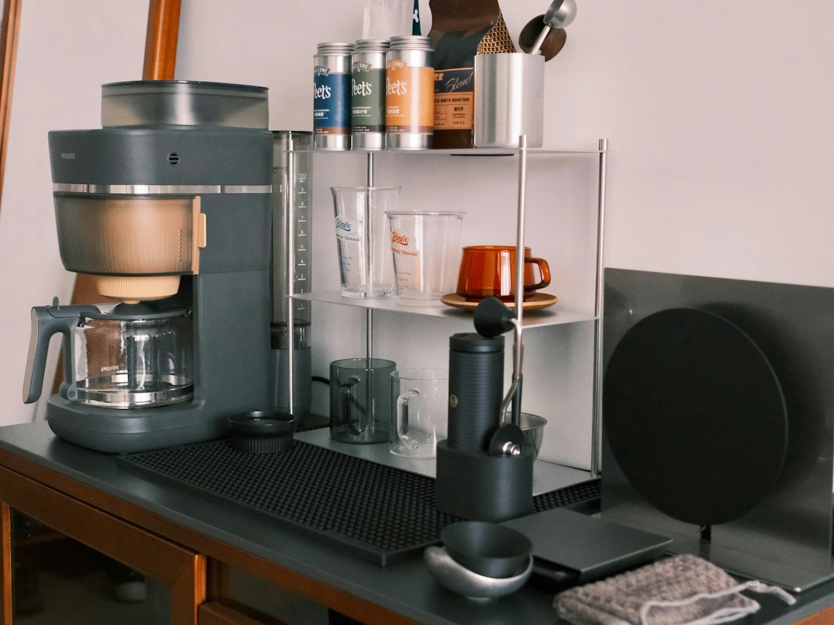 A compact kitchen counter coffee station with a drip coffee maker, a hand grinder, glass measuring cups and jars of coffee beans on a small shelf unit.