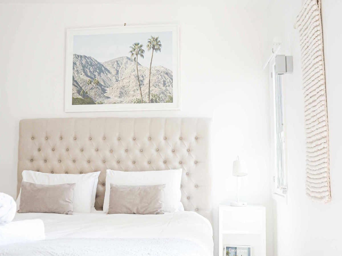 A neatly made bed with a beige tufted headboard and white and taupe pillows, in a bright bedroom.