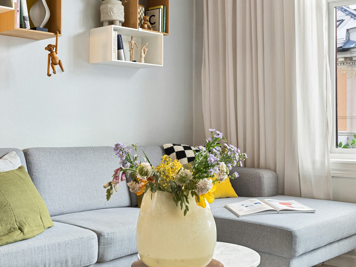 A grey sofa and chaise with cushions and a vase of wildflowers, with wall-mounted shelves and sheer curtains at a window.