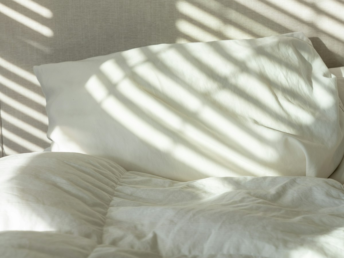 Close-up of rumpled white bedding and a pillow, with sunlight casting blind-slat shadows across the fabric.