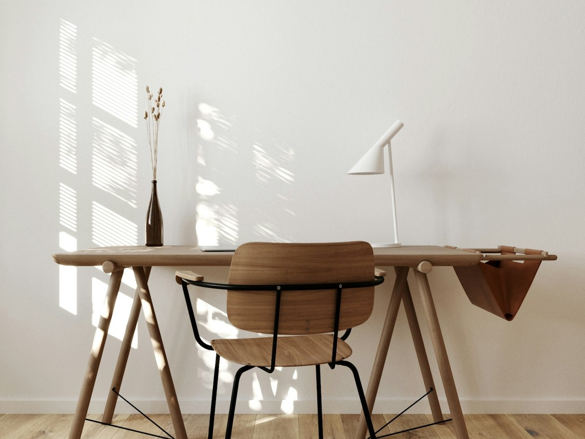 A pale wood trestle desk with a wooden chair and a white adjustable desk lamp, lit by slatted window light.