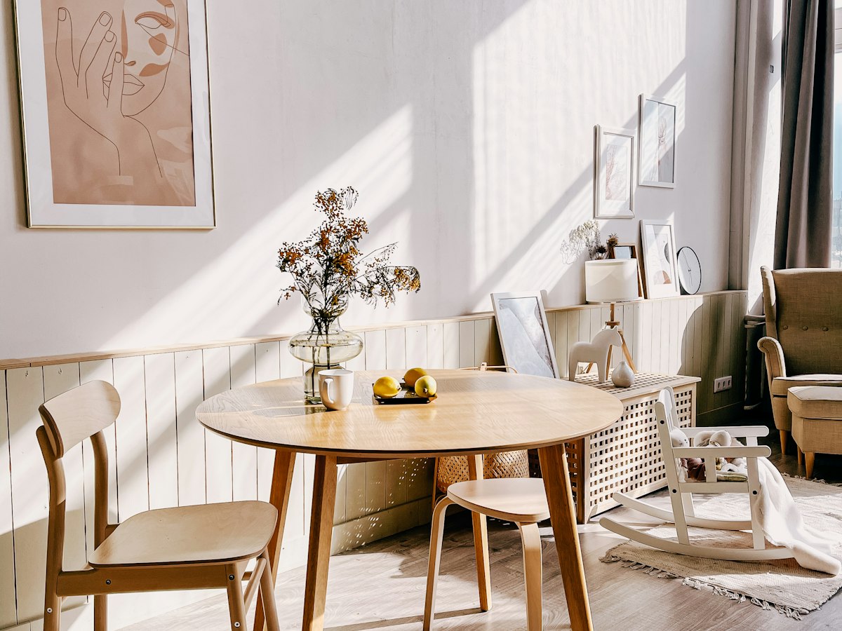 A small round wooden dining table with two wooden chairs and a rocking chair nearby, beside a sunlit window.