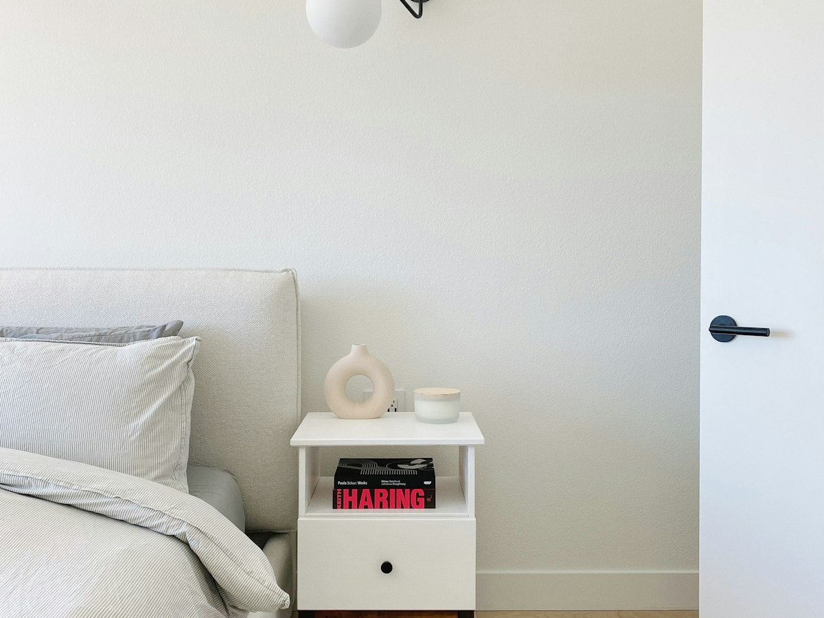 A bed beside a small white bedside table holding books, a candle and a ceramic vase, in a minimal bedroom.